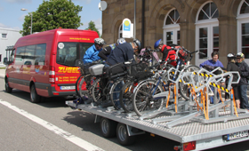 Mehrere Personen verladen ihre Fahrräder auf einem speziellen Anhänger, der an einem roten Bus angehängt ist.